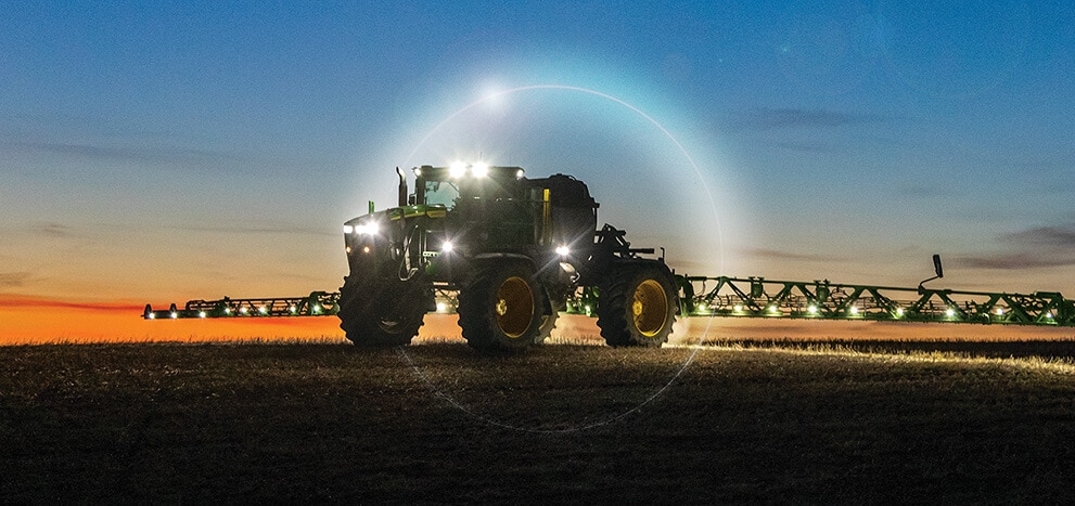 Self-propelled sprayer with extended boom operating in a field at dusk, headlights illuminated.