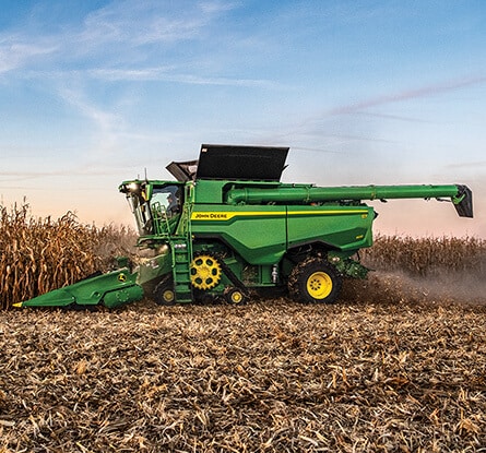 Green combine harvesting corn in a field, leaving dust behind under a clear sky.