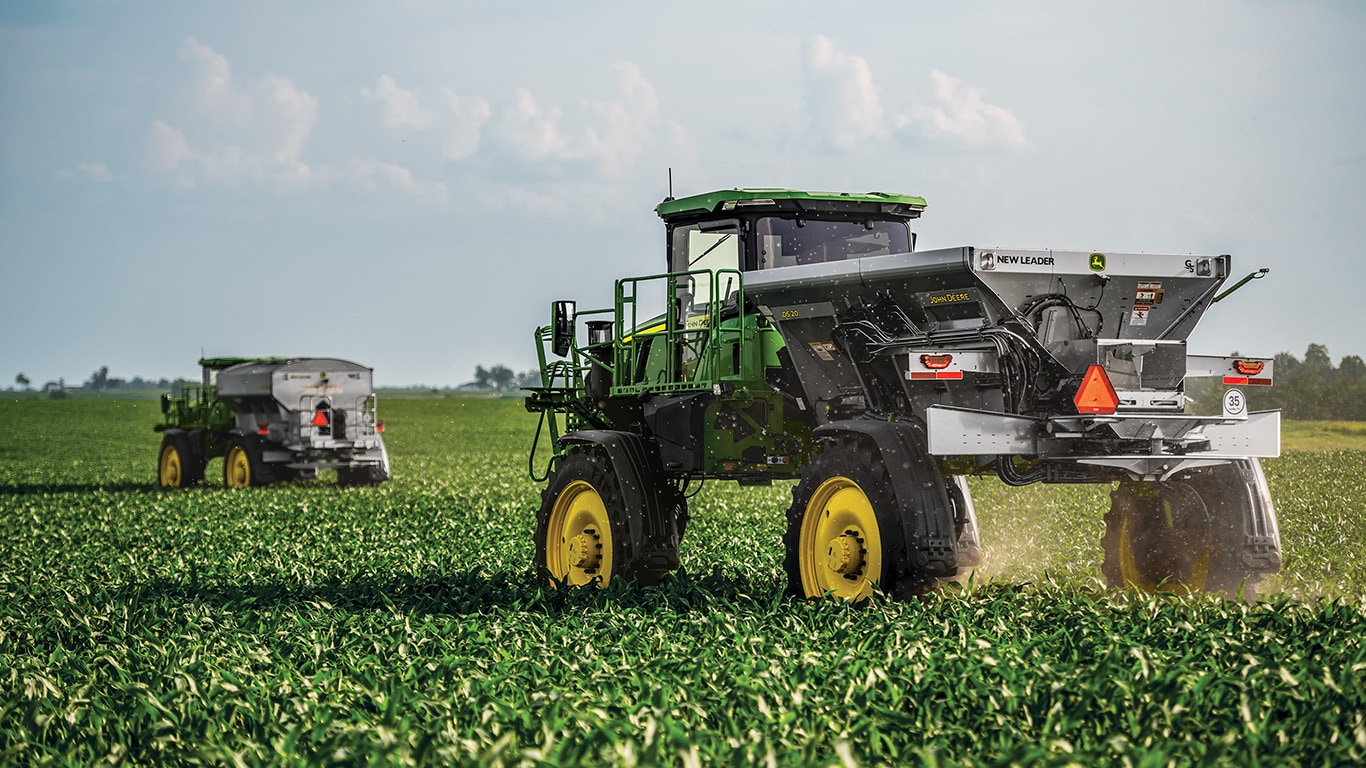 Two John&nbsp;Deere self-propelled spreaders operating in a green agricultural field, with the foreground machine dispersing material and a second spreader visible in the distance under a partly cloudy sky.