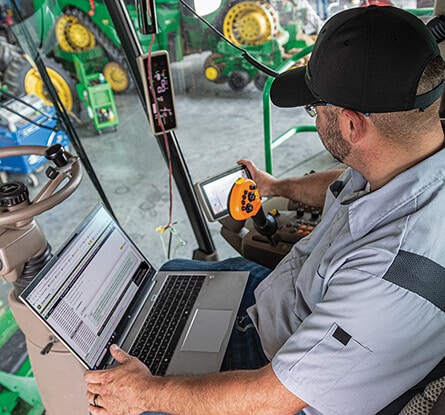 Person inside tractor cab using a laptop and touchscreen monitor for equipment diagnostics.