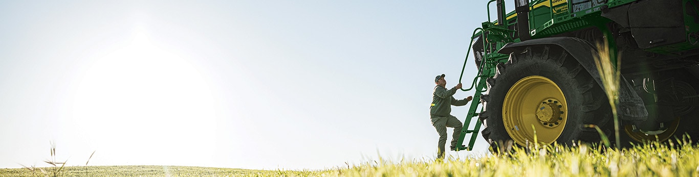 Person climbing the steps of a large John&nbsp;Deere tractor parked in a grassy field under a bright sky.