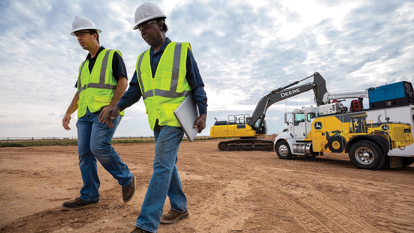 Operators walking across a worksite with a John Deere excavator and work truck in the background. 