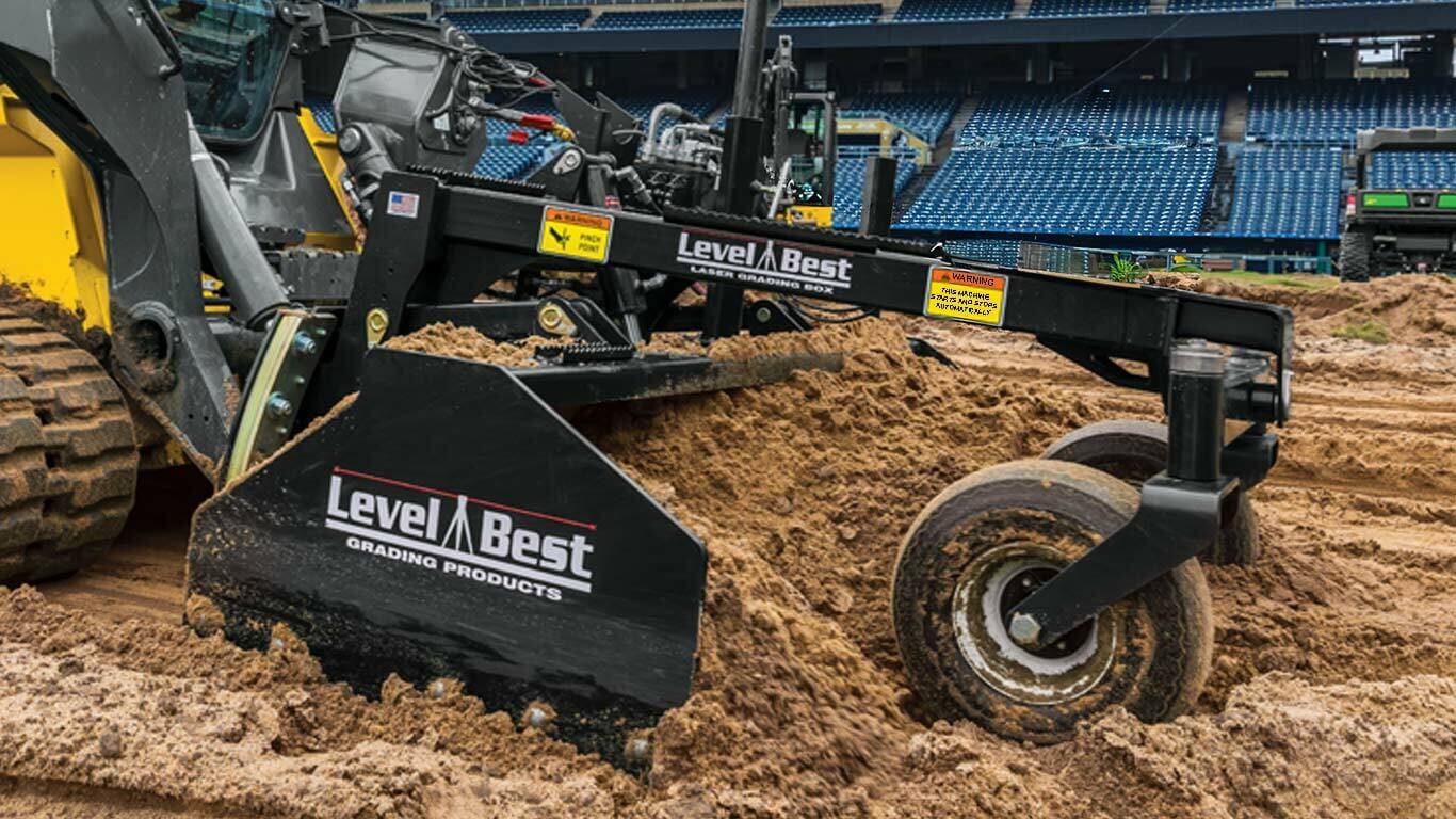 A Level Best grading machine smoothing dirt on a construction site with stadium seating in the background.