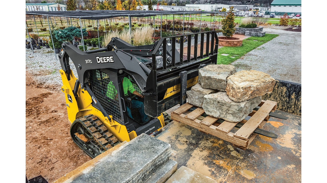 A 317G Compact Track Loader with pallet forks attachment lifting a pallet of large rocks at a nursery.