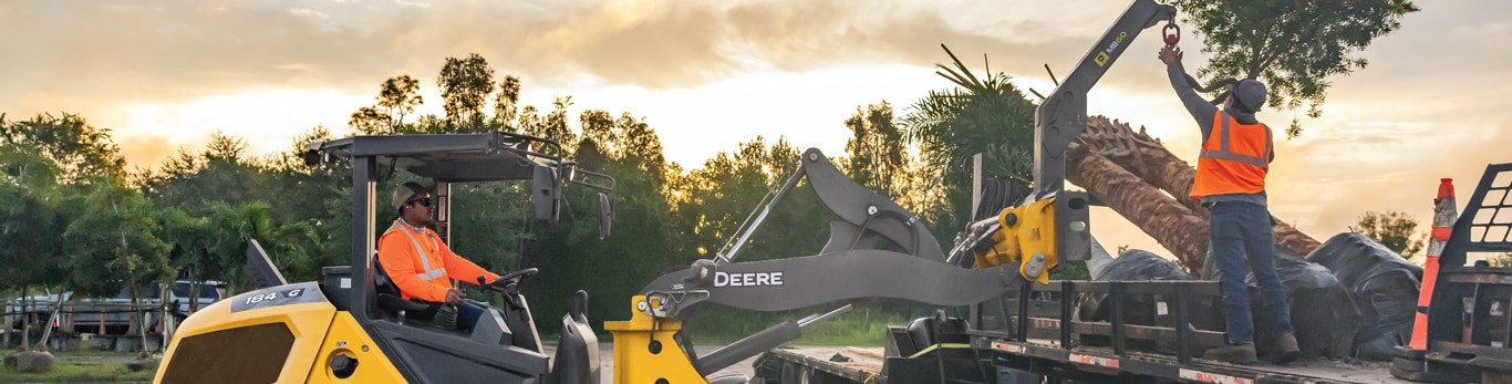 A worker in a John Deere 184 G-Tier Compact Wheel Loader lowers a MB60 attachment to pick up a palm tree.