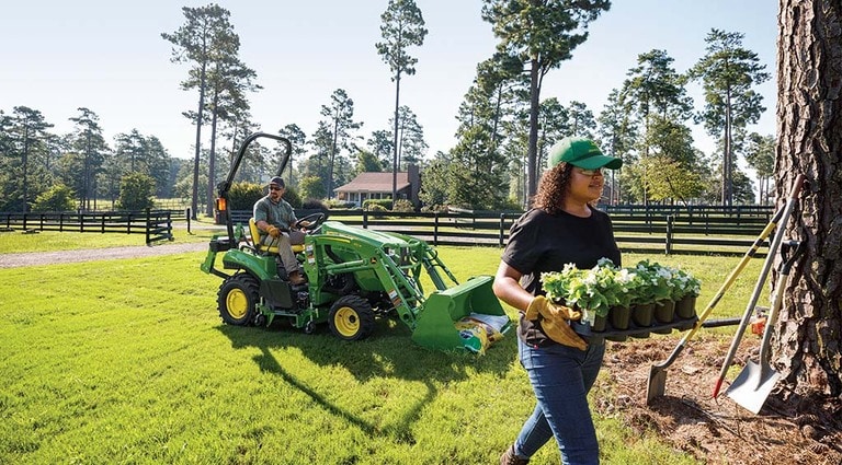 A person carrying plants, a 1023E Tractor with soil bags is resting behind them.