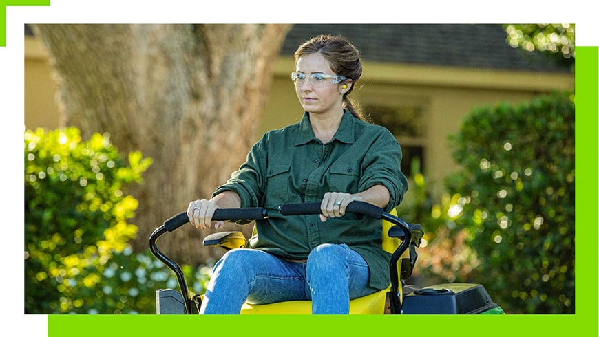 A woman with dark brown hair and a dark green shirt and jeans on rides a green and yellow John Deere Z370R Electric lawn mower with safety glasses on.