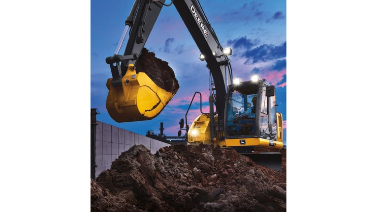 A 135P-Tier Excavator with headlights scooping dirt from a pile at a worksite with sunset in the background.