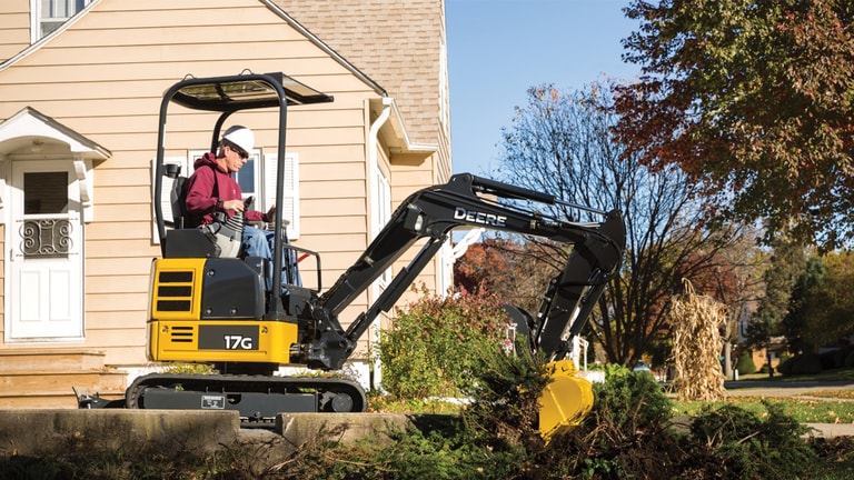 Man using 17G excavator to dig up a bush in front of a house.
