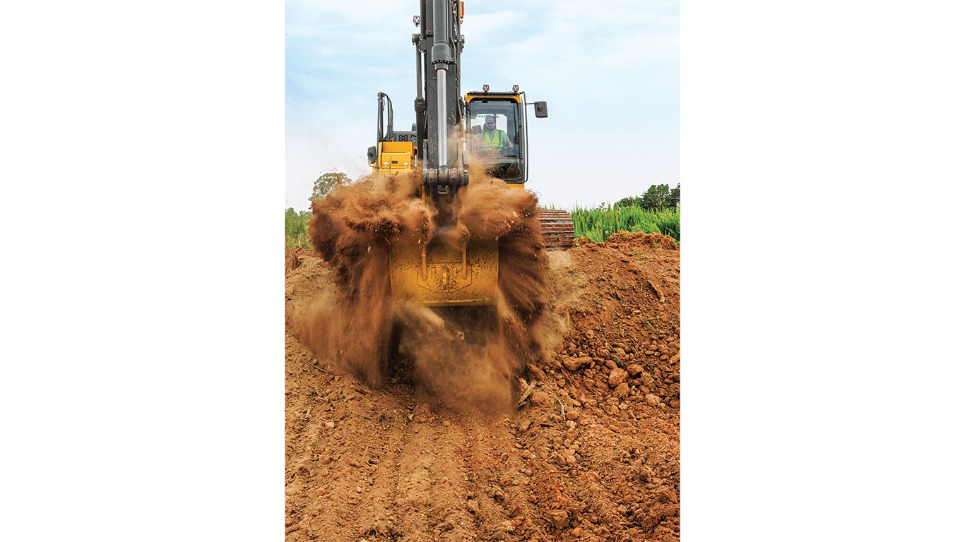 A front-facing perspective of an operator scooping up dirt with a 210P-Tier Excavator with a green field in the background.