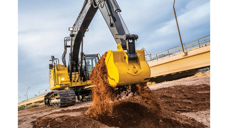 An operator using a 245 P-Tier Excavator to scoop dirt at a worksite with an overpass bridge in the background.