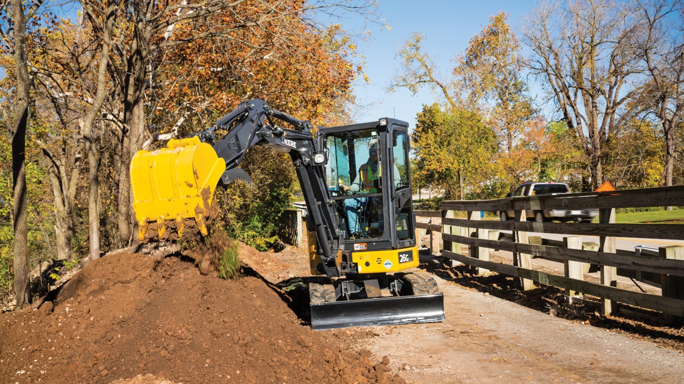 26G Excavator dropping topsoil into a stockpile on a sidewalk in front of a wooden fence.