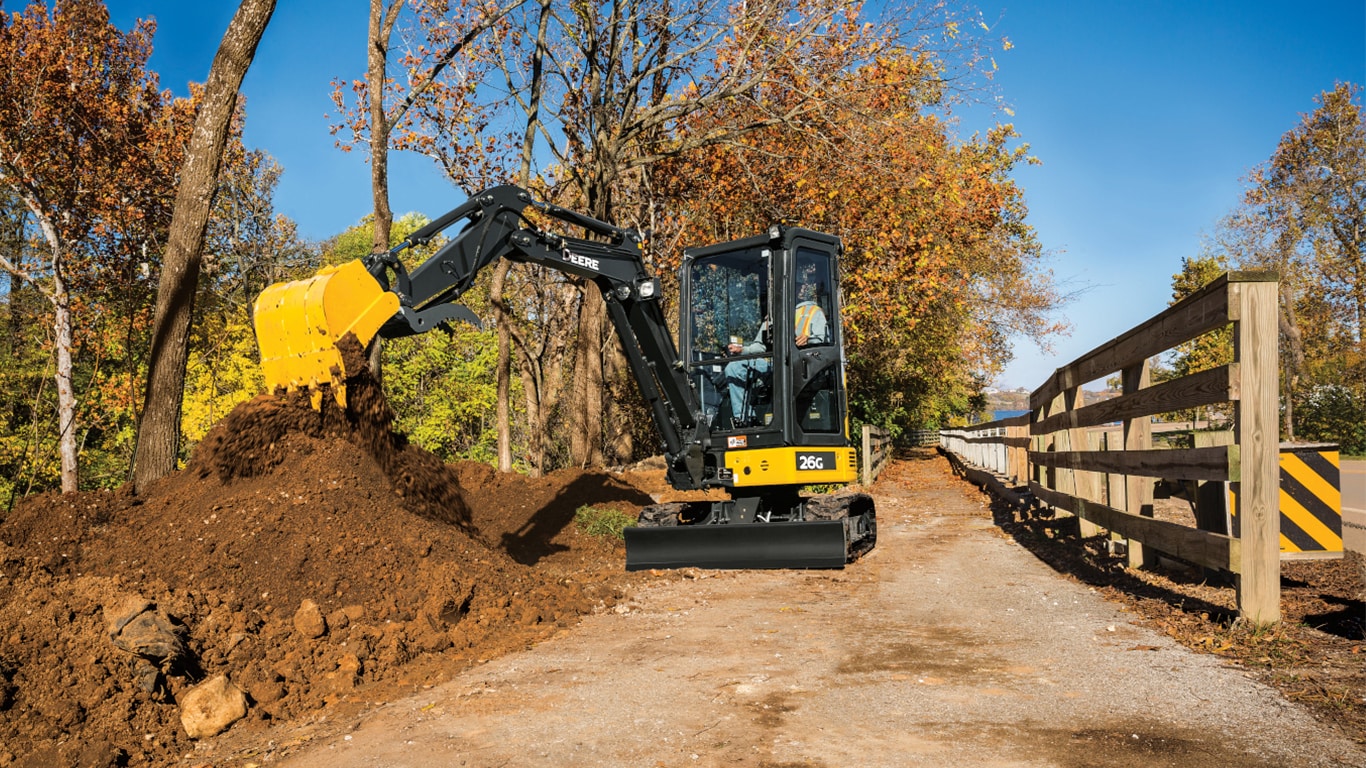 Man using 26G Excavator to stockpile dirt on a sidewalk with a wooden fence.