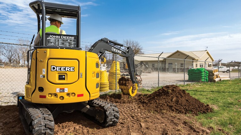 An operator moving topsoil to the right of a John Deere 30G Excavator in front of a chain link fence.