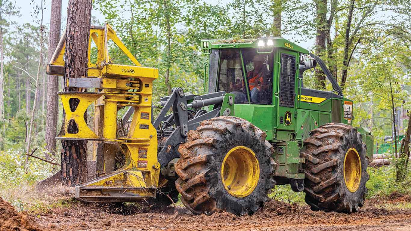 Green wheeled feller buncher gripping a tree trunk with a yellow cutting head in a forest.