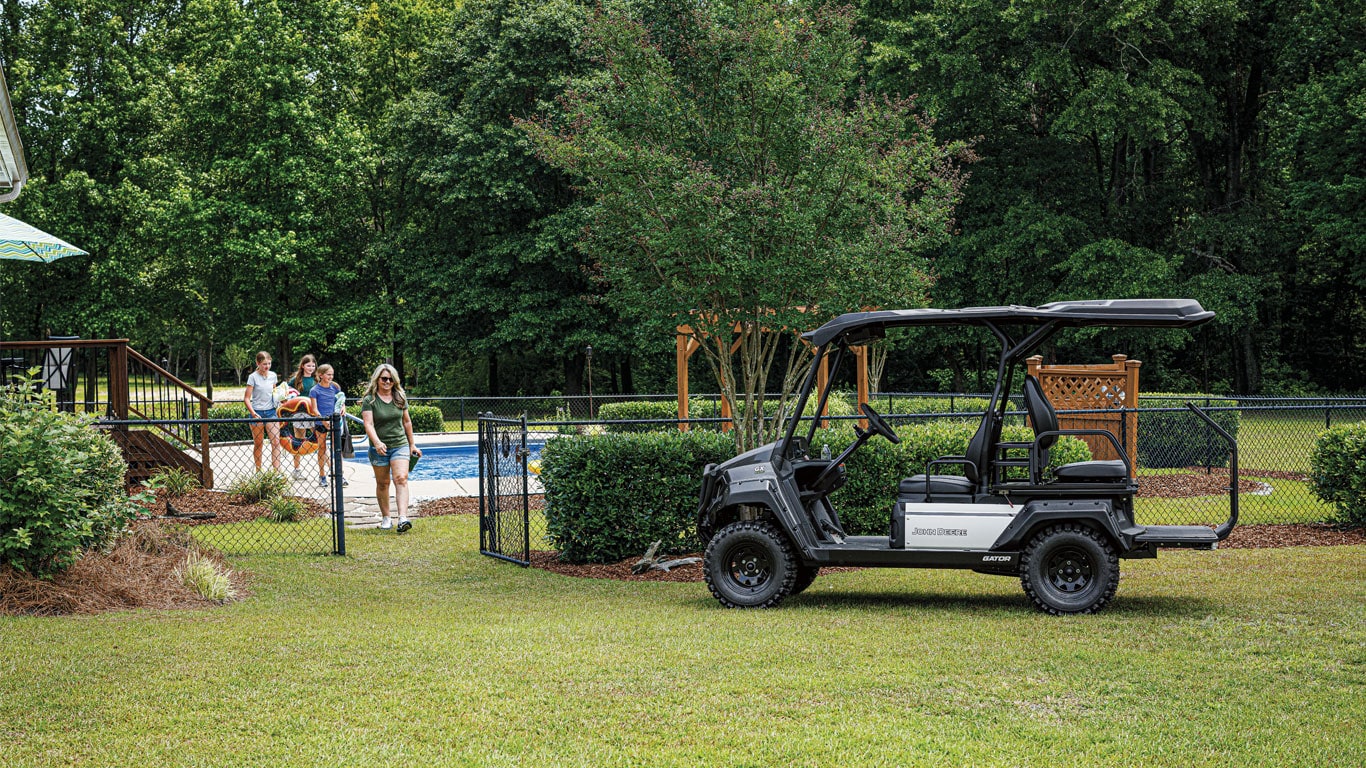 The new John Deere GX electric Gator parked as a family exits a fenced in pool area, headed towards the vehicle