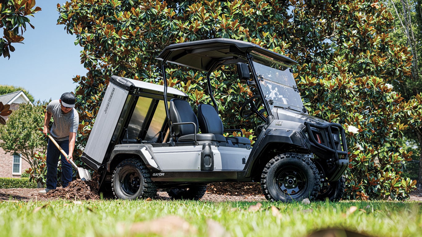 Ground level shot of the new John Deere GX electric Gator with the cargo bed tilted down as a property owner use a shovel to remove wood chips from the bed and make a pile