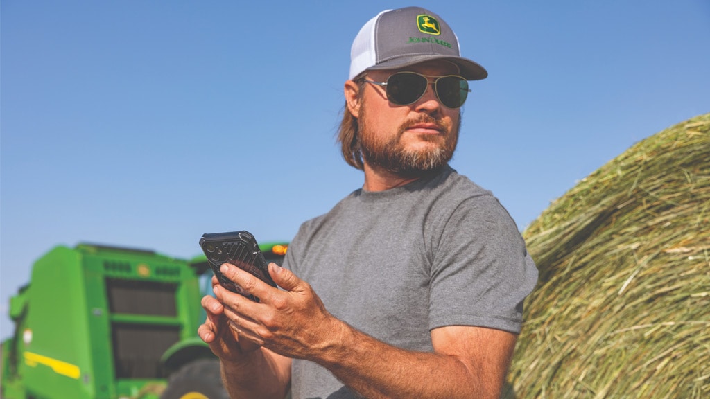 Person leaning on round bale with cell phone in hand and a 1 Series Round Baler in the background