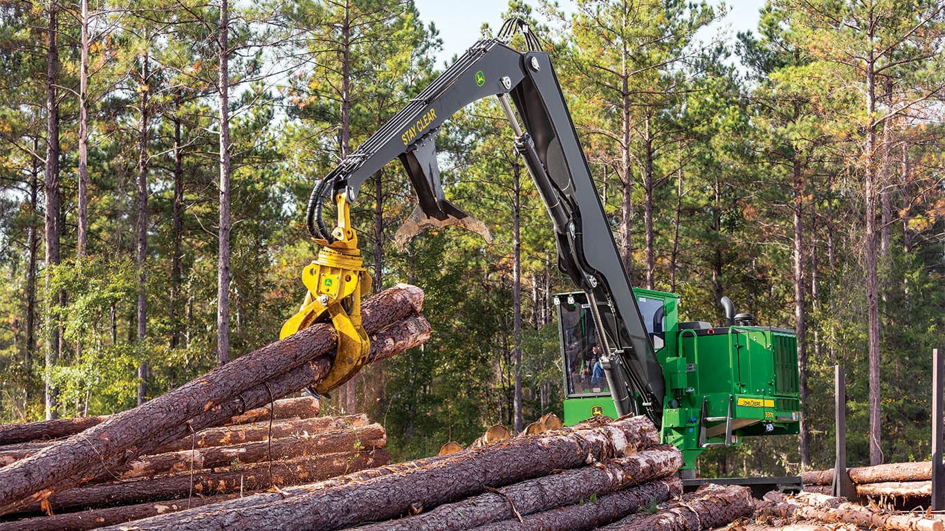 A John&nbsp;Deere knuckleboom loader adding logs to a pile of logs in a forest. 