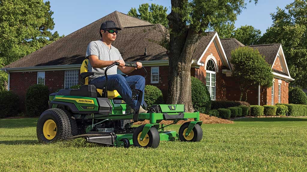 A person with a cap and sunglasses driving a green electric zero-turn mower around a residence.