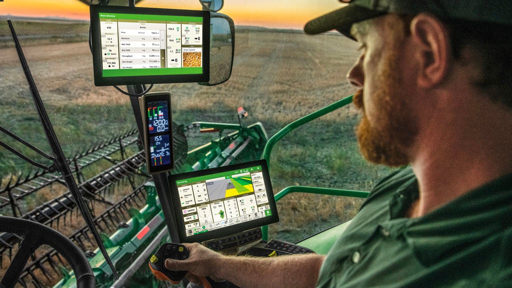 In-Cab photo of a farmer driving a John Deere Combine in a field at sunset. He's looking at a illuminated G5Plus Display. He also has a G5Plus Extended Monitor above a Primary Corner Display.