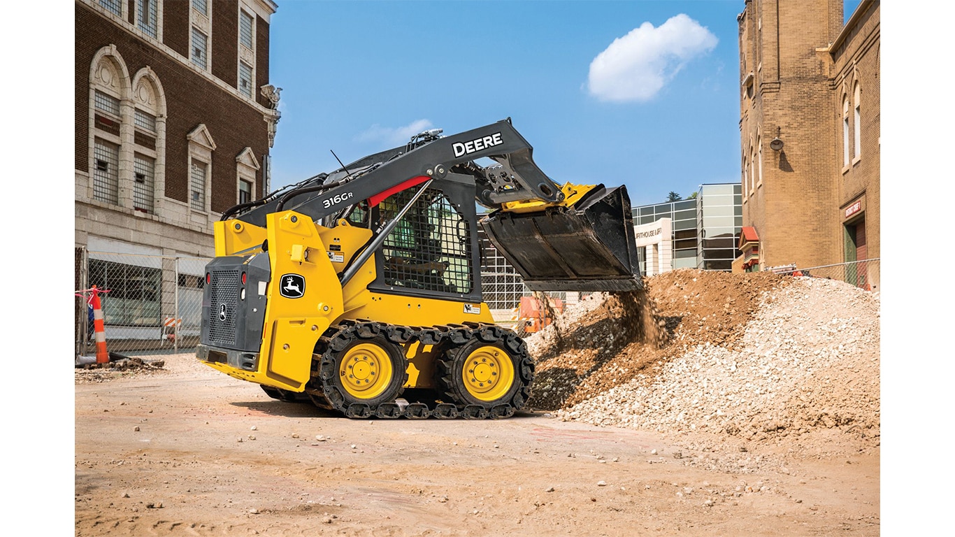 Un Minicargador 316G descarga rocas y tierra en una pila en un lugar de trabajo con edificios en el fondo.