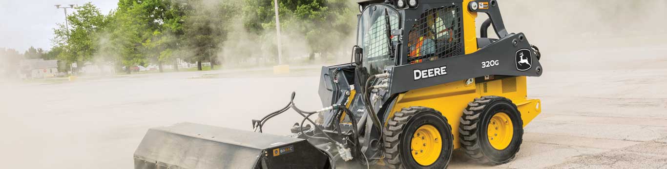 320G Skid Steer with angle broom attachment sweeps a parking lot in a cloud of dust