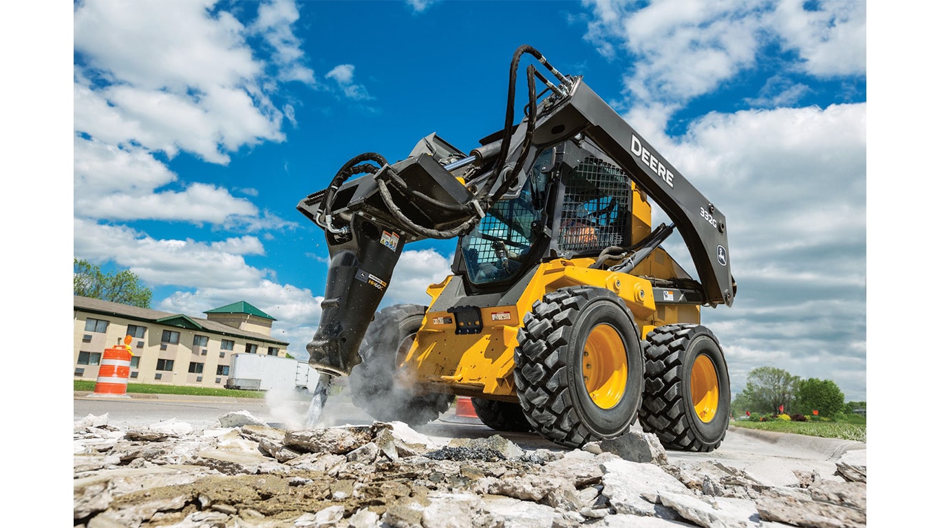 A 332G Skid Steer with a hydraulic hammer attachment breaking up concrete in front of a building.