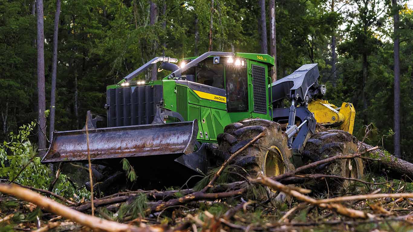 Green skidder with blade and grapple clearing logs in a dense forest.