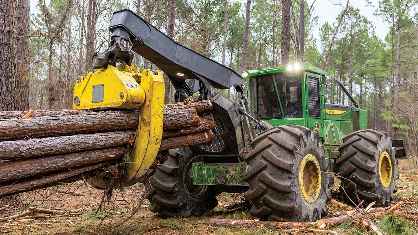 Green skidder lifting several logs in a forest clearing with large rugged tires.