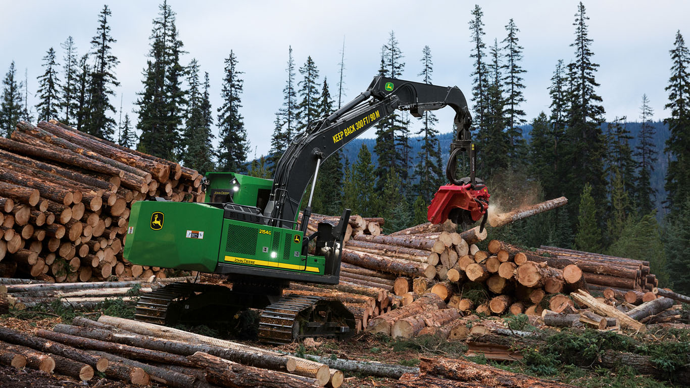 A John&nbsp;Deere swing machine dropping logs onto a pile with trees in the background.