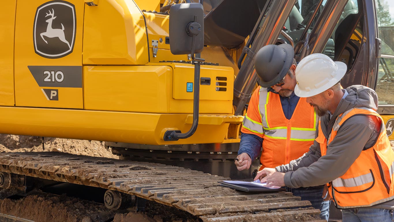 Two construction workers in safety gear review documents on a clipboard beside a yellow excavator.
