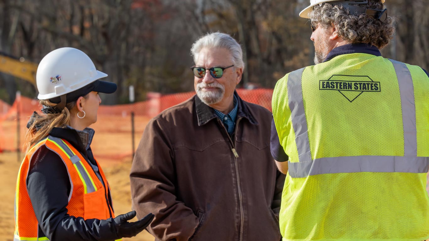 Three people in safety gear converse at a construction site with orange fencing and trees in the background.
