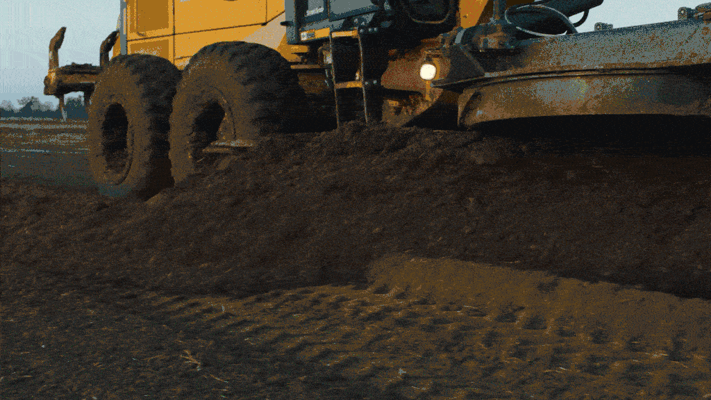 A yellow construction vehicle with large tires is grading and moving soil on a dirt road.