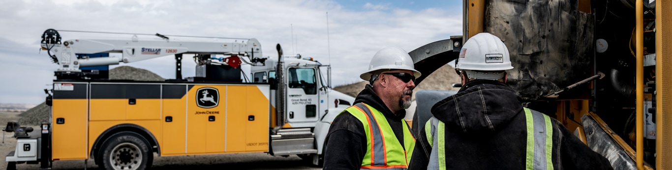 Dealer technicians work on a piece of construction equipment at a jobsite with a service truck in the background.