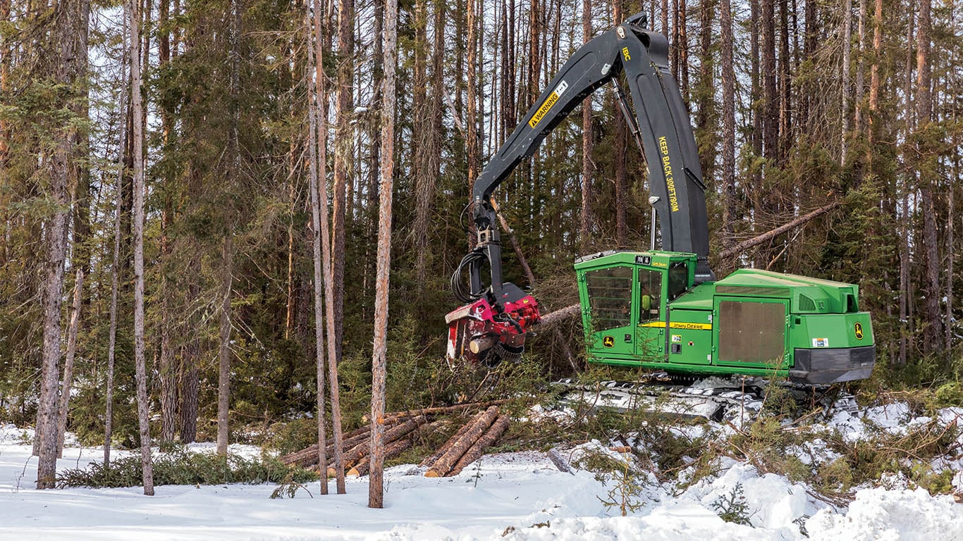A John Deere tracked harvester cutting logs to length in a snowy forest.