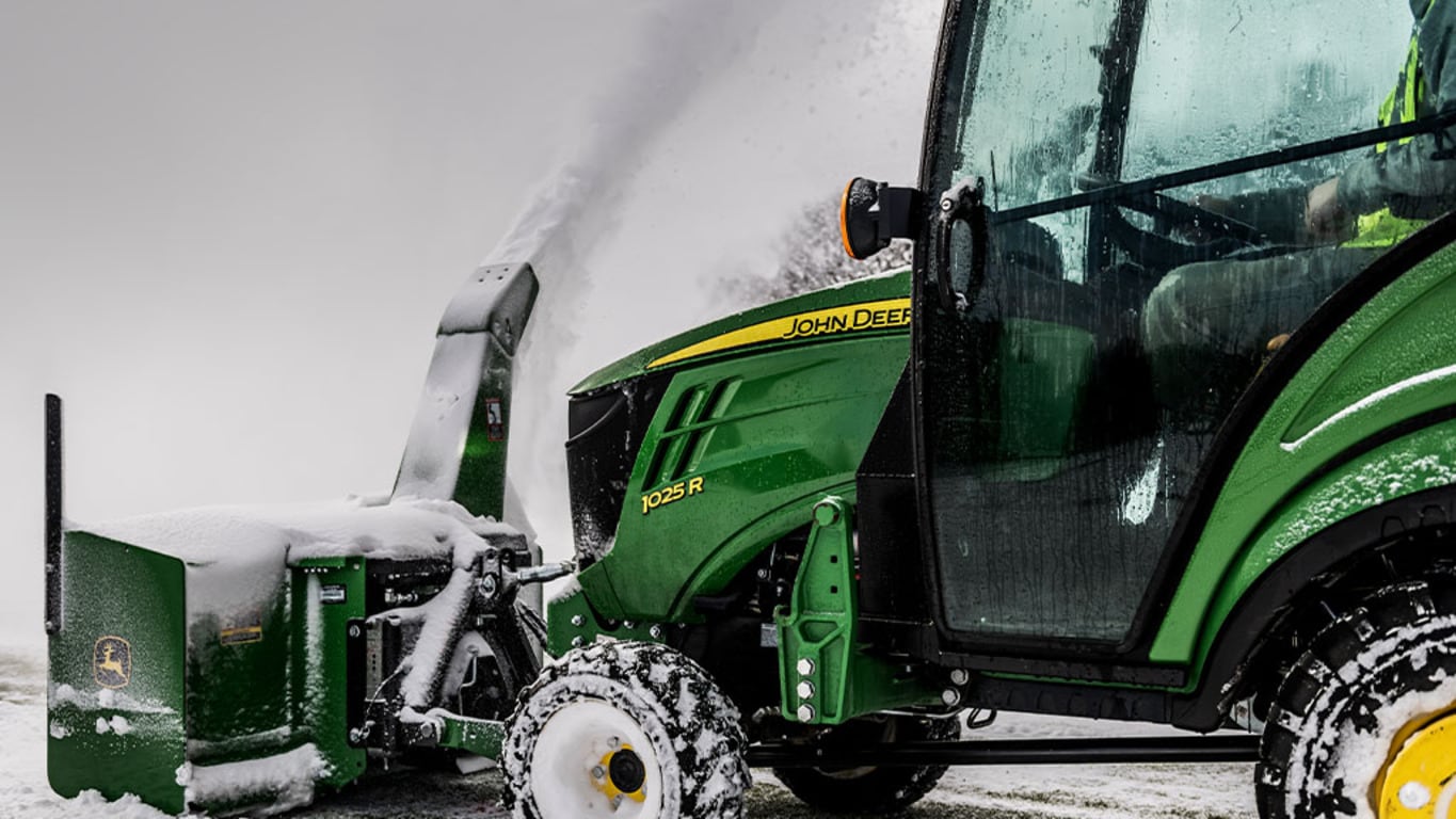 A green compact tractor with a front-attached snow-blower removing snow from a path.