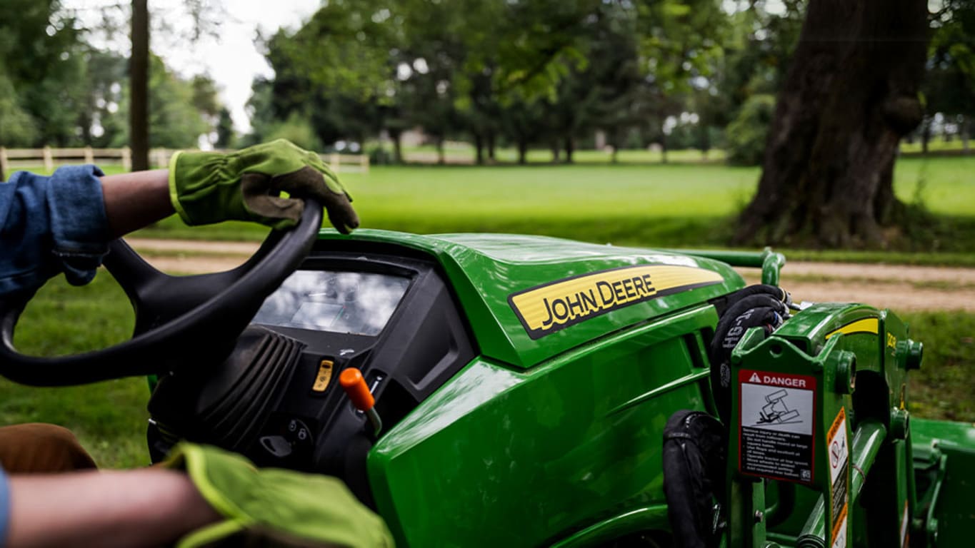 Close up of the John Deere logo on the front hood of a compact tractor while a gloved hand rest on the steering wheel.