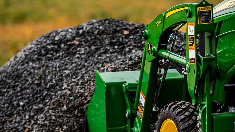 A green front loader attached to a compact tractor digs into a pile of gravel ready to lift it up.