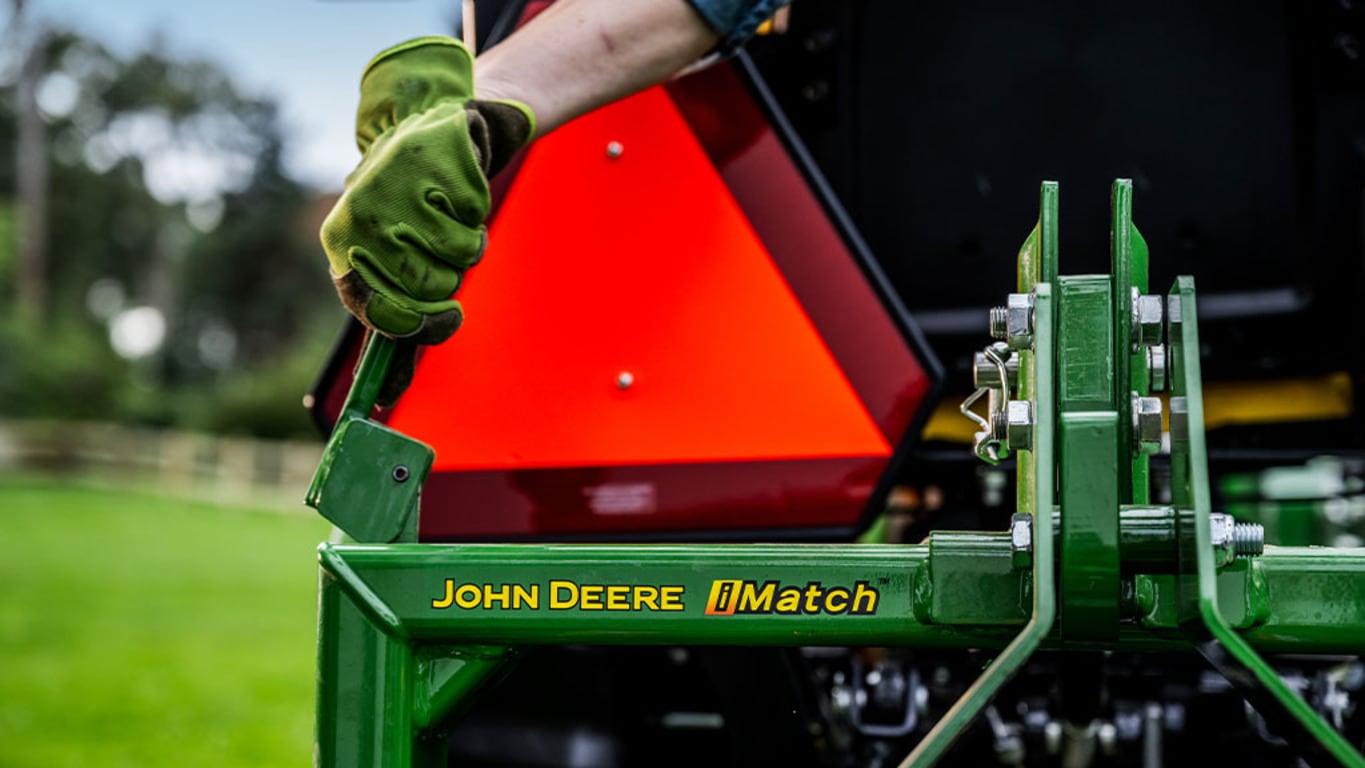 A gloved hand pulls a lever in front of the rear SMV emblem near the John Deere iMatch logo.