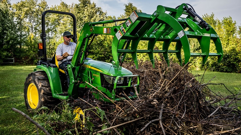 A debris grapple attached to a green compact tractor is open about to grab a pile of branches.