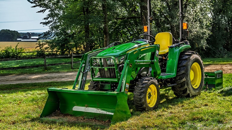 A person approaches a green compact tractor resting in the sun with a loader and box blade attached to it.