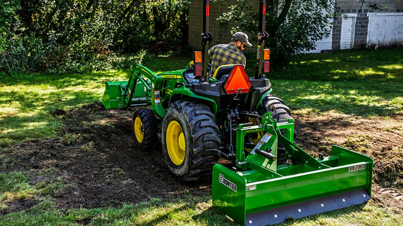 A front-loader and box blade attached to a green compact tractor scrapes off the top soil and grass to create a dirt patch.