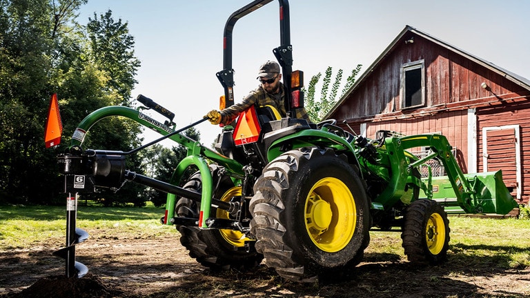 A post-hole digger on the back of a green compact tractor is creating a hole near a red barn.
