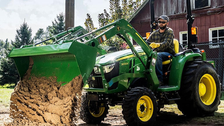 Dirt is dumped in the middle of four buried posts by a front loader attached to a green compact tractor.