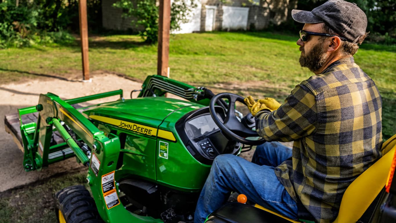 A pallet of pavers is carried to a cleared dirt patch by a green compact tractor with a pallet fork attachment.