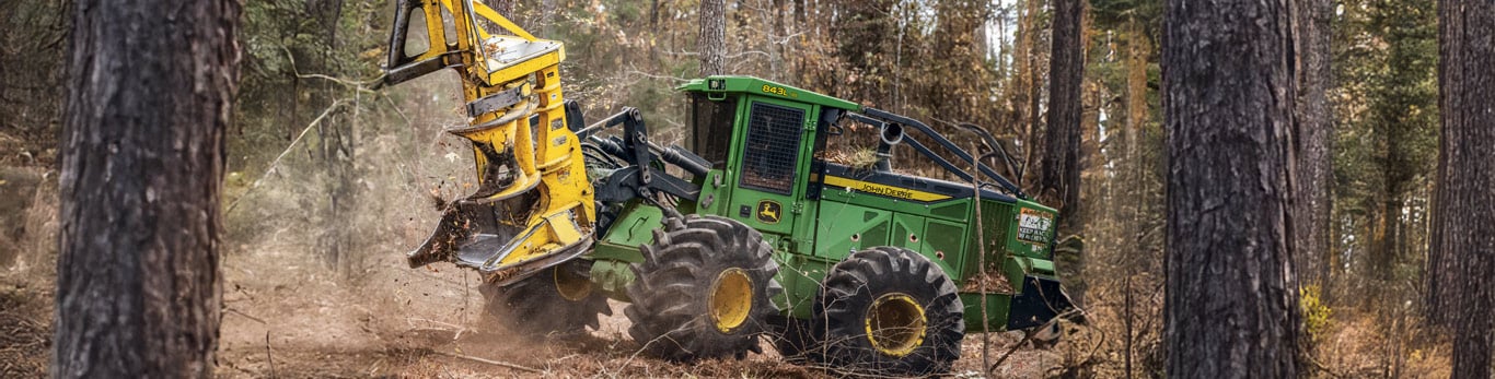 A John Deere 843L-III Wheeled Feller Buncher driving through a forest.