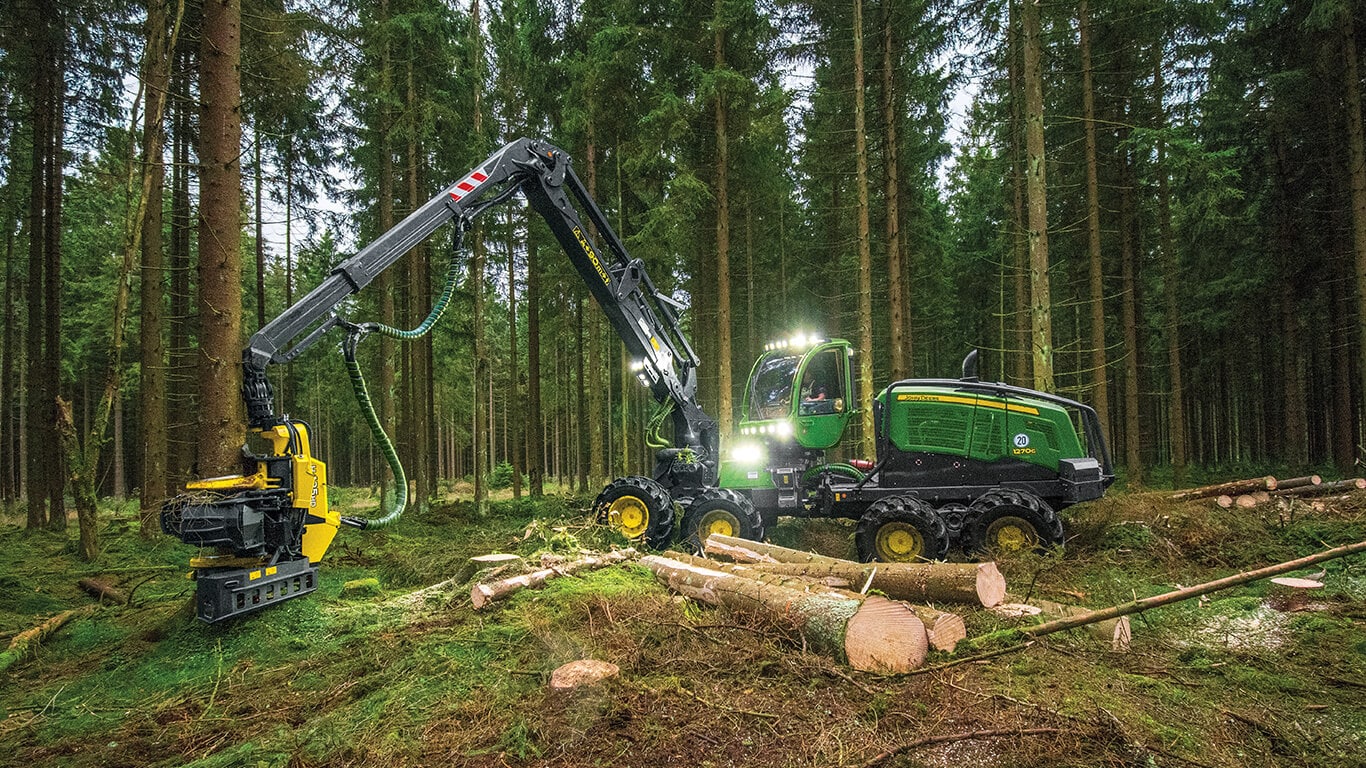 John&nbsp;Deere wheeled harvester working in wooded area with lights on harvesting trees.