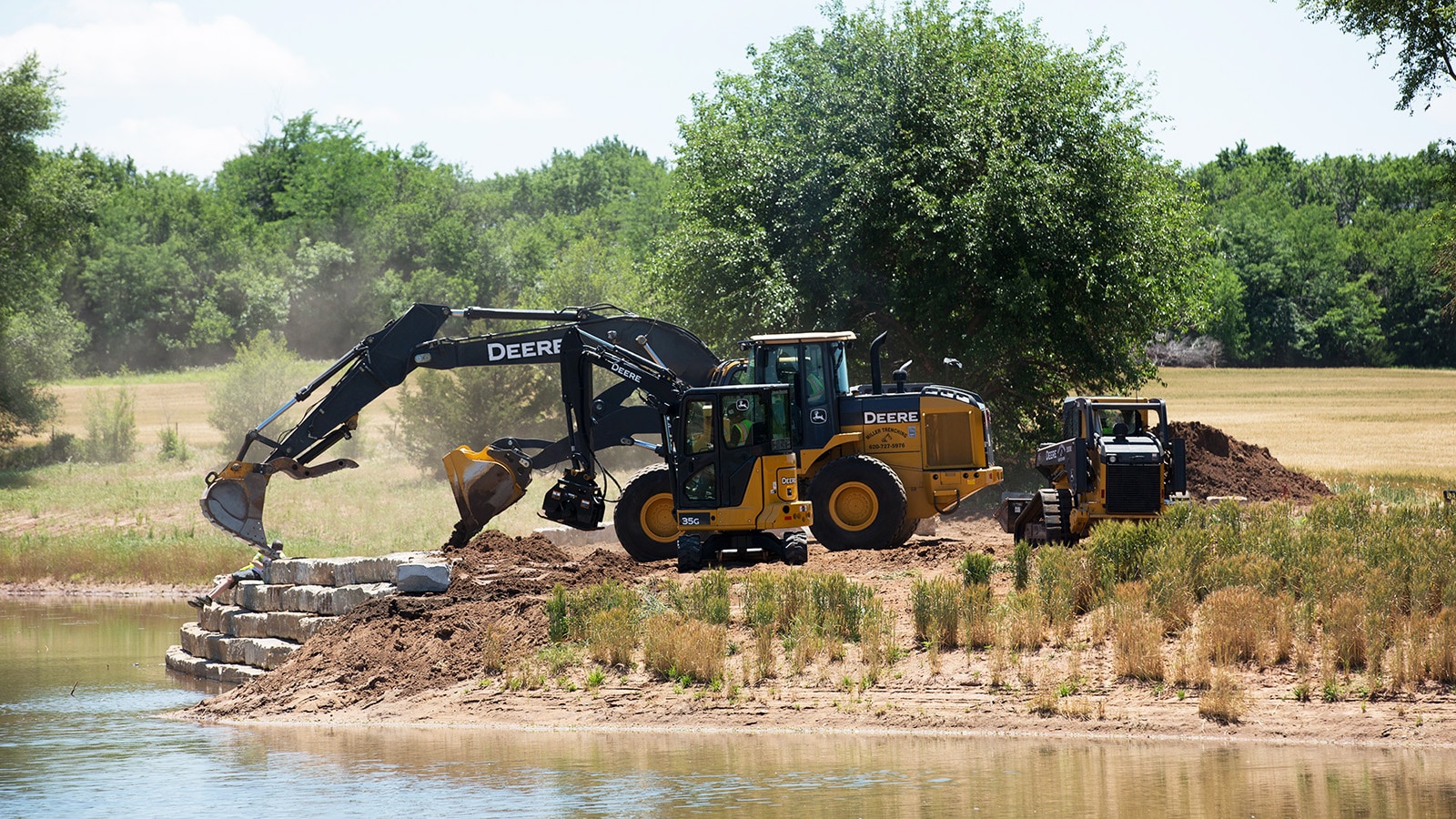 Wheel loader, excavators and compact track loaders work on a retaining wall by a pond.