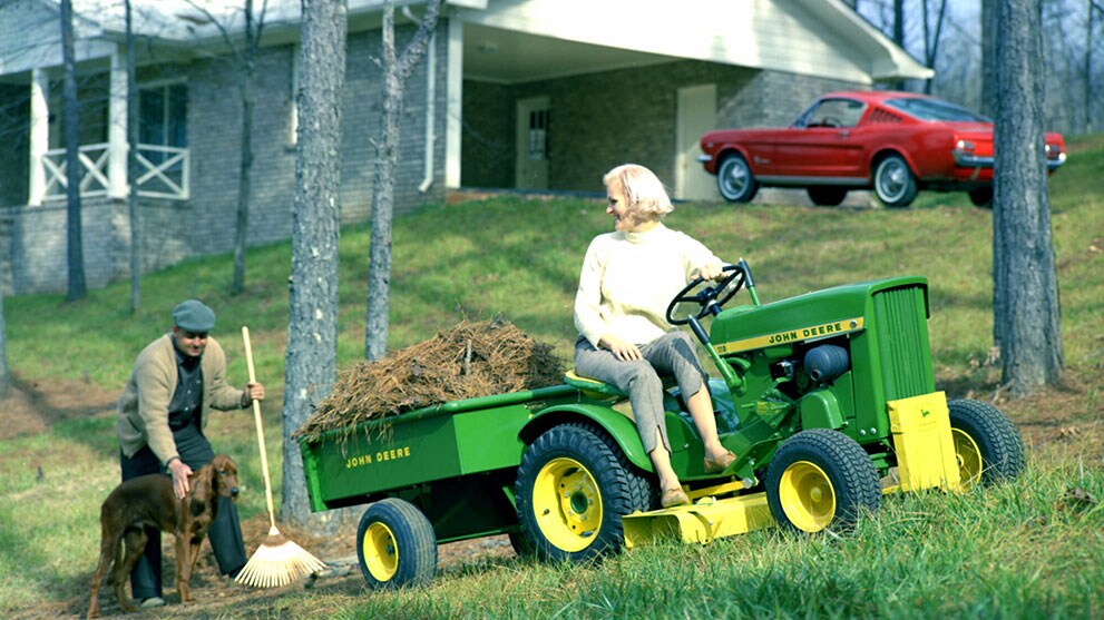 A woman operates an older model lawn tractor with a full dump cart trailing behind